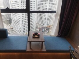 A room with a view of tall buildings through a window, a small table with a decorative item, and a blue cushioned bench.