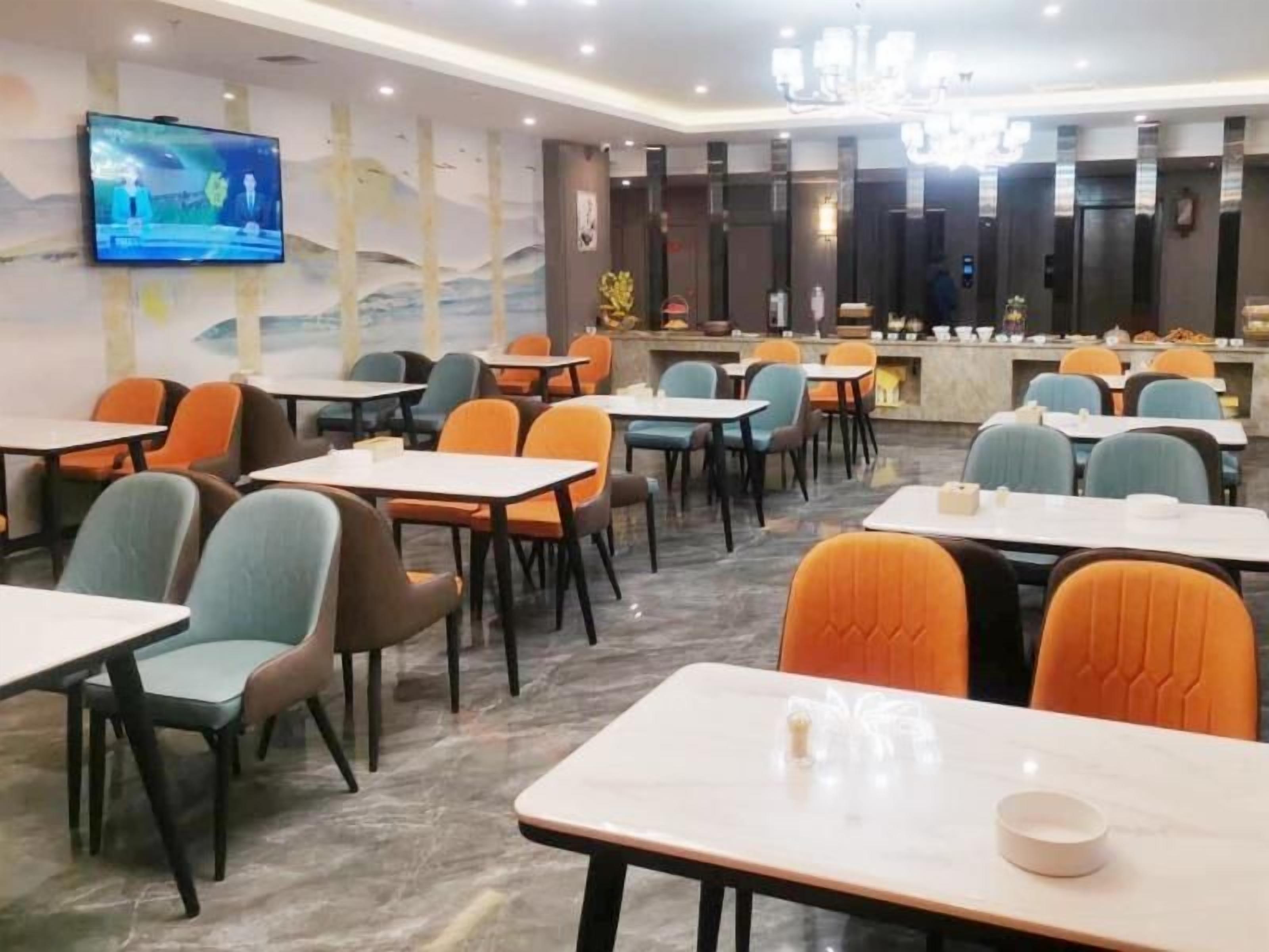 A hotel dining area with multiple tables and chairs, a television mounted on the wall, and a reception desk in the background.