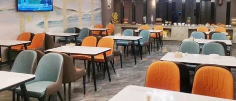 En hotelrestaurant med flere borde og stole, et fjernsyn ophængt på væggen og en reception i baggrunden.