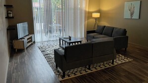 A living room with a sofa, a coffee table, a TV, and a lamp. There is a sliding glass door leading to an outdoor area.