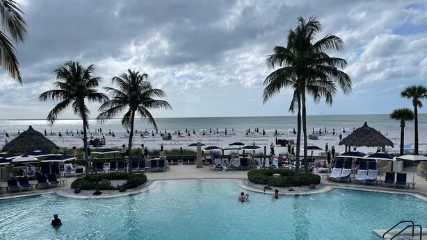 A pool area with lounge chairs and umbrellas, surrounded by palm trees and a beach in the background.
