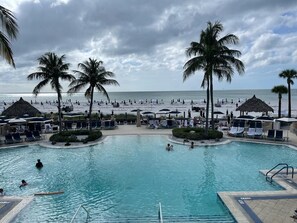 Ein Poolbereich mit Liegestühlen und Sonnenschirmen, umgeben von Palmen und einem Strand im Hintergrund.
