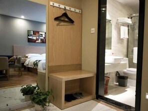 A hotel room with a bed, a wooden shelf, a shoe rack, a potted plant, and a bathroom visible through a glass door.