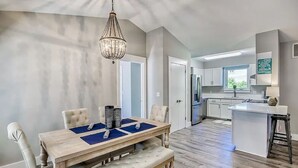 A modern kitchen with a central island, a dining area with a wooden table and cushioned chairs, and a chandelier.