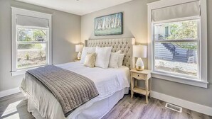 A bedroom with a large bed, two bedside tables with lamps, a ceiling fan, and a window with a view of a staircase and greenery.