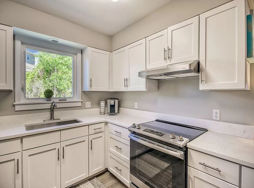A modern kitchen with white cabinets, a stainless steel range hood, a built-in oven, and a window with a view of greenery.