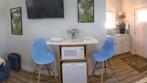 Un espace de vie compact comprenant un coin cuisine, un coin repas avec deux chaises bleues, un téléviseur à écran plat et des œuvres d’art décoratives murales.