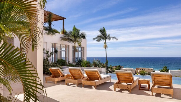 A poolside area with wooden lounge chairs, a view of the ocean, and a building with large windows. - Topia Sunset Suite All Inclusive (El Pescadero)