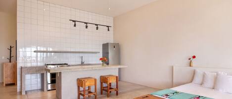 A modern kitchen with a central island, bar stools, a refrigerator, and a bed with a colorful blanket.