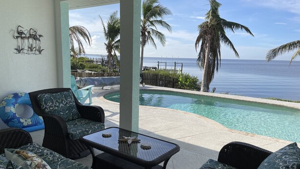 A poolside area with wicker furniture and a glass table, overlooking a clear blue ocean and palm trees.