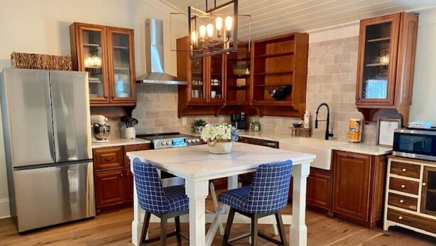 Una cocina con armarios de madera, un refrigerador de acero inoxidable, una mesa de comedor blanca con sillas azules y una lámpara colgante.