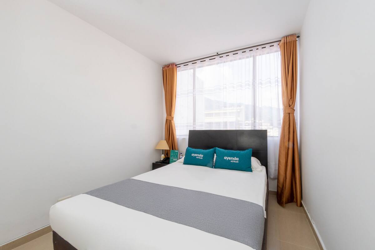 A hotel room with a large bed, two pillows with the brand name, a nightstand with a lamp, and a window with curtains.