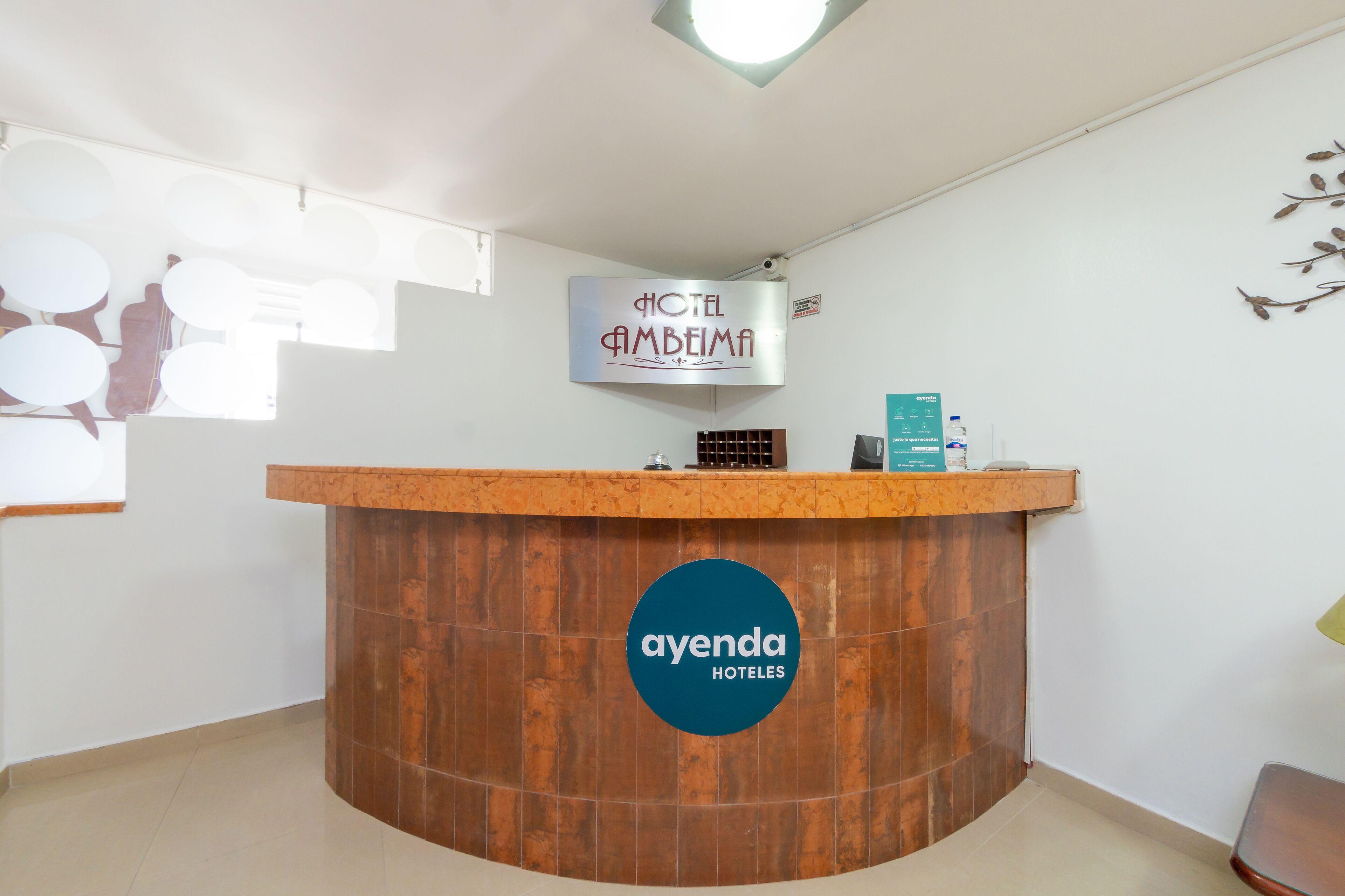 A hotel reception desk with a wooden finish and a blue logo. A sign above the desk reads "HOTEL AMBEIMAH".