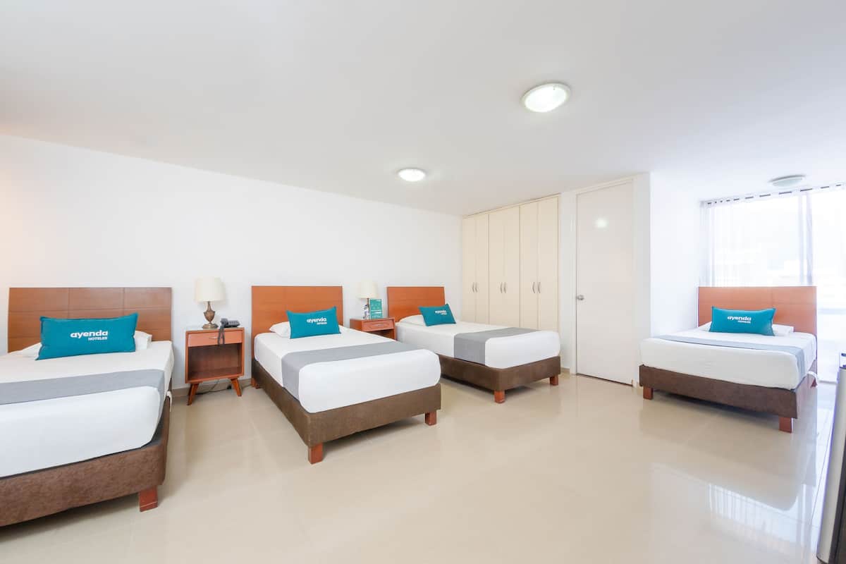 A hotel room with three beds, each with a wooden headboard and a blue pillow with the hotel name. There are bedside tables with lamps and a small table between the beds. The room has a large wardrobe and a window with blinds.
