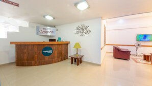 A hotel reception area with a wooden reception desk, a small table with a lamp, a wall-mounted TV, and a massage chair. - Ayenda Ambeima (Ibagué)