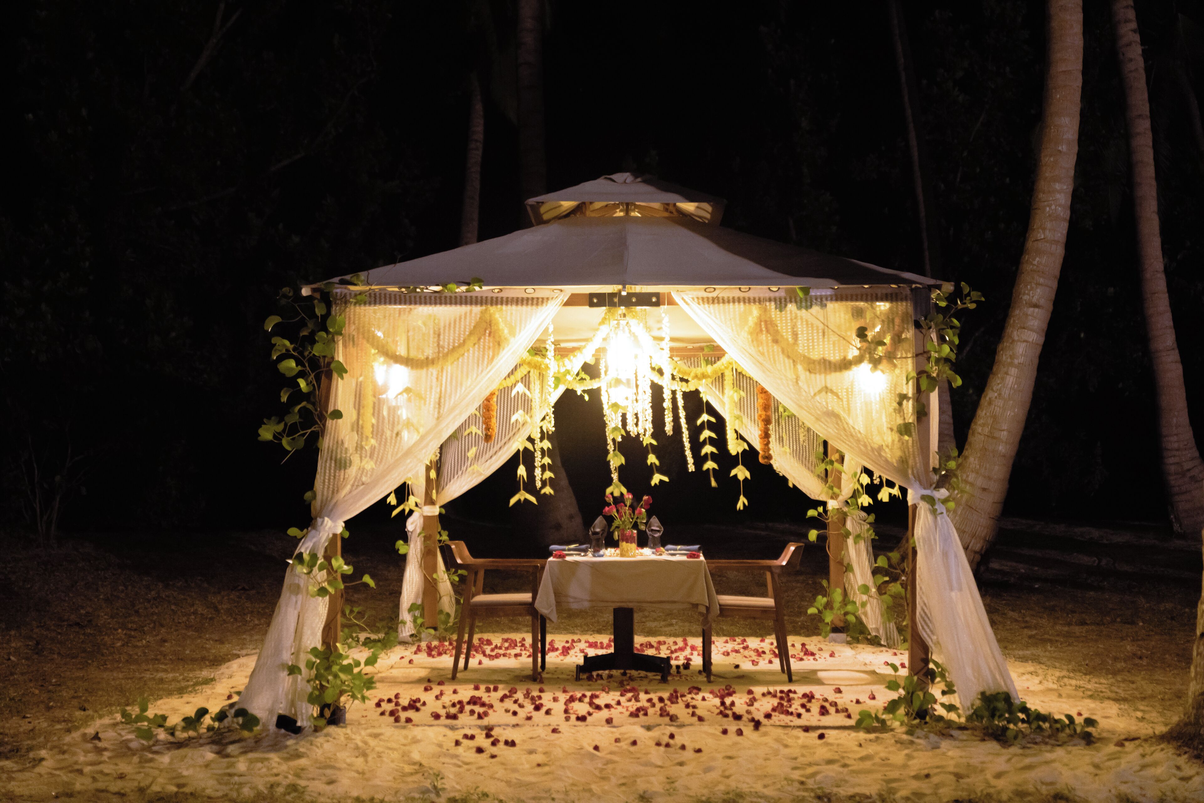 Un área de comedor al aire libre con toldo, con una mesa preparada para dos personas, rodeada de arena y pétalos de rosa dispersos.