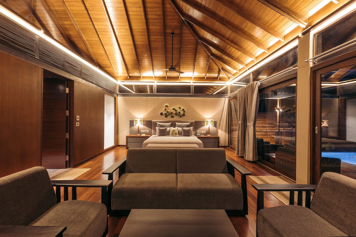 Quarto de hotel moderno com teto de madeira, uma cama grande, área de estar e vista para uma piscina.