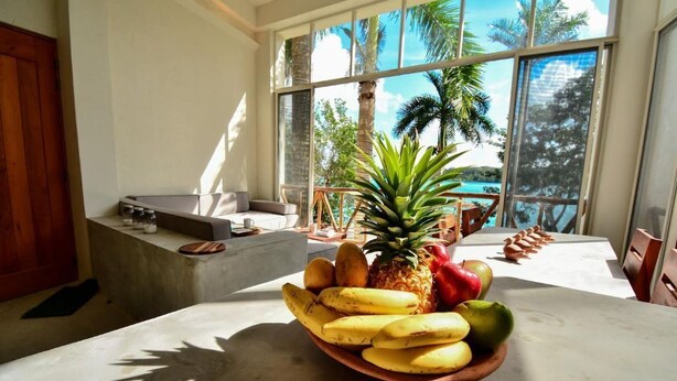A dining area with a fruit bowl on a table, large windows with a view of palm trees, and a concrete bench. - Villa IV Casa Kuuts Ja (Casa de patos) (Xul-Ha)