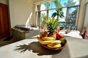 Een eetgedeelte met een fruitschaal op een tafel, grote ramen met uitzicht op palmbomen, en een betonnen bank.