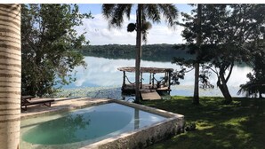 A serene outdoor pool area with a wooden deck and lounge chairs, surrounded by lush greenery and a calm lake. - Villa IV Casa Kuuts Ja (Casa de patos) (Xul-Ha)