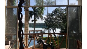 A room with a large window overlooking a lake, palm trees, and a wooden deck. - Villa IV Casa Kuuts Ja (Casa de patos) (Xul-Ha)