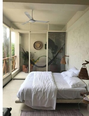 A bedroom with a bed, a ceiling fan, a hammock, and a view of greenery outside.