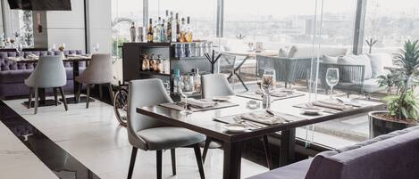 Un espace repas moderne avec une longue table, des chaises et un canapé. On y trouve également un coin bar avec des bouteilles et des verres.
