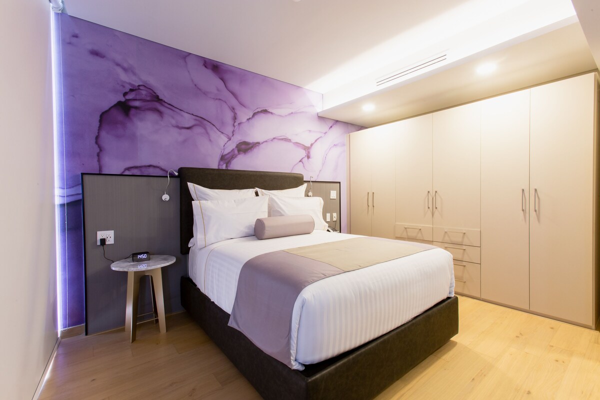 Une chambre à coucher moderne avec un grand lit, une table de chevet, une armoire et un mur décoré d’un motif imitant le marbre.