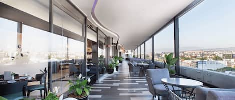 Un área de descanso moderna con asientos y mesas, amplias ventanas con vistas a la ciudad y un diseño de techo curvo.
