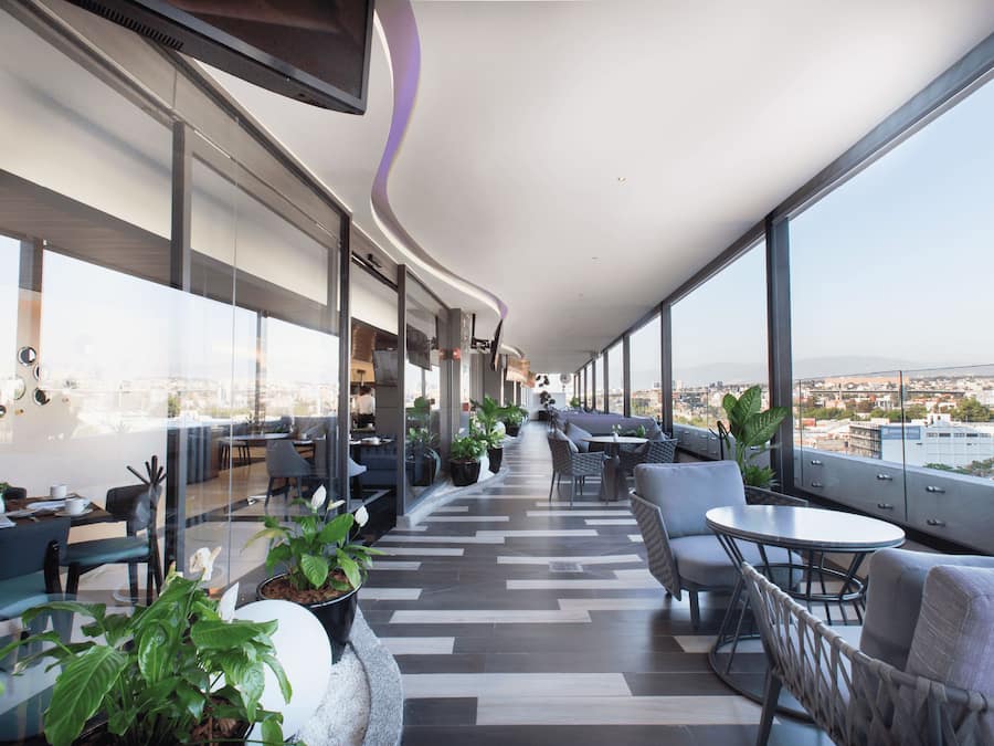 Un área de descanso moderna con asientos y mesas, amplias ventanas con vistas a la ciudad y un diseño de techo curvo.