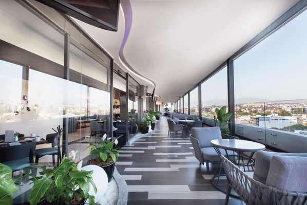 Un área de descanso moderna con asientos y mesas, amplias ventanas con vistas a la ciudad y un diseño de techo curvo.