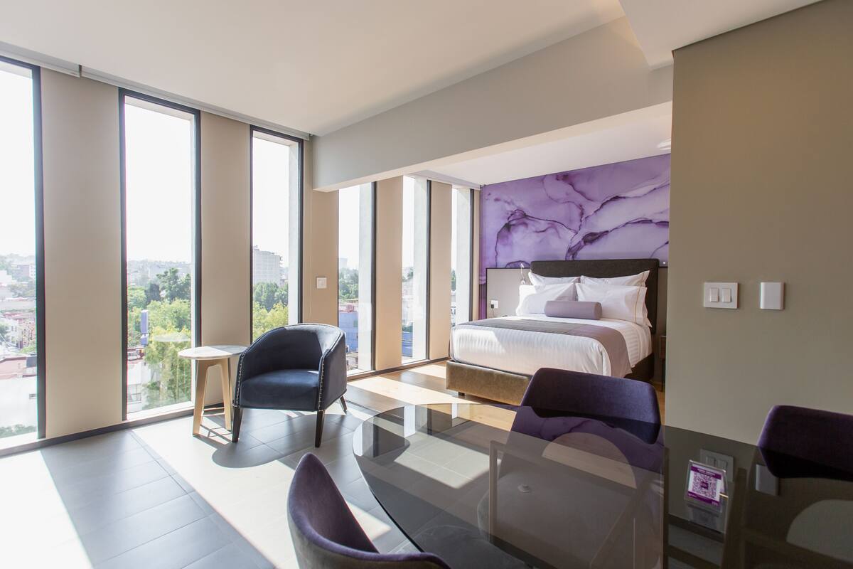 Een moderne hotelkamer met een groot bed, een glazen tafel, twee stoelen en uitzicht op de stad door grote ramen.
