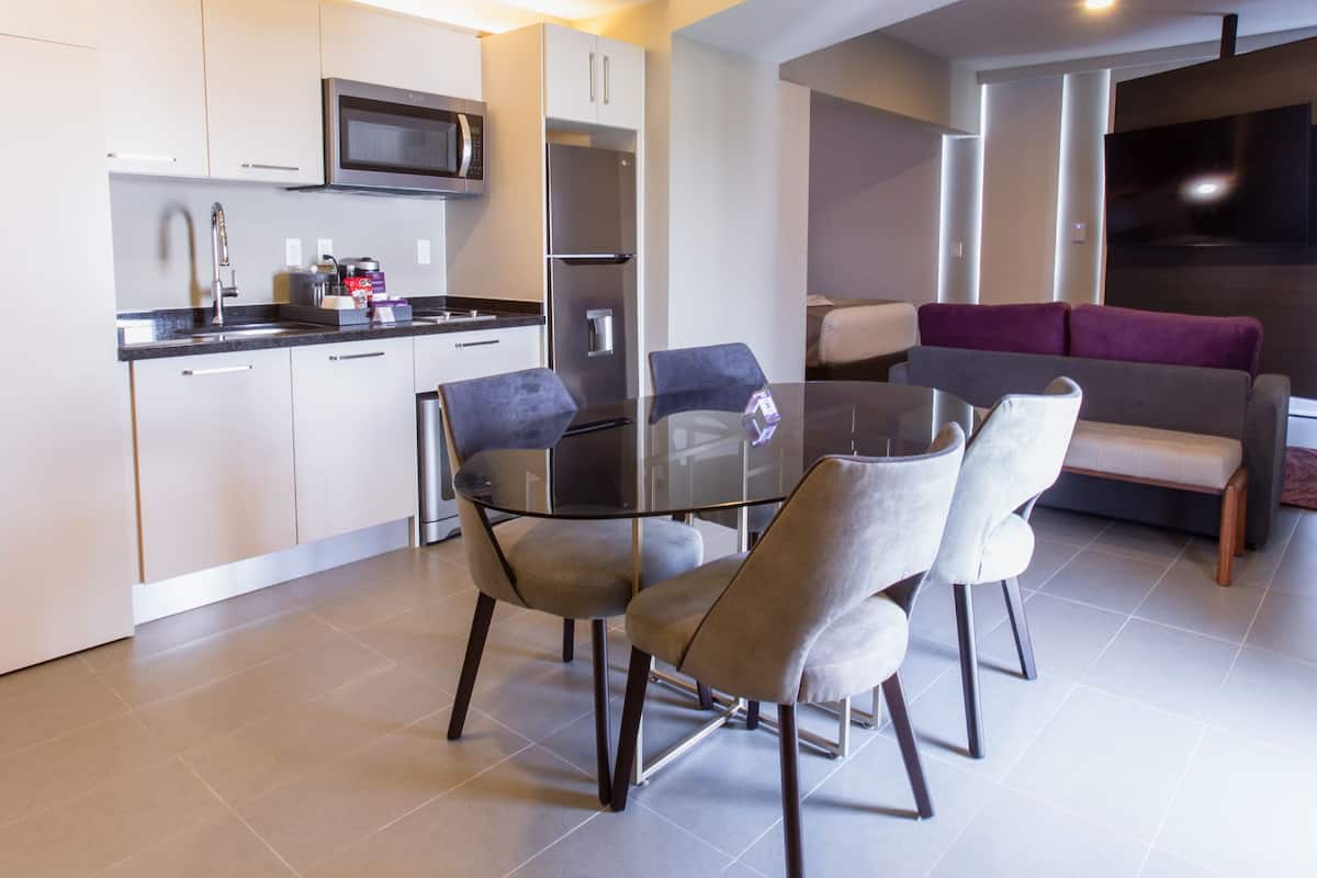 Une cuisine moderne avec un coin repas comprenant une table et des chaises en verre, ainsi qu’un salon avec un canapé et un téléviseur.
