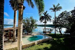 En swimmingpool omgivet af palmer og en pavillon, med udsigt til en sø og fjerne bakker.