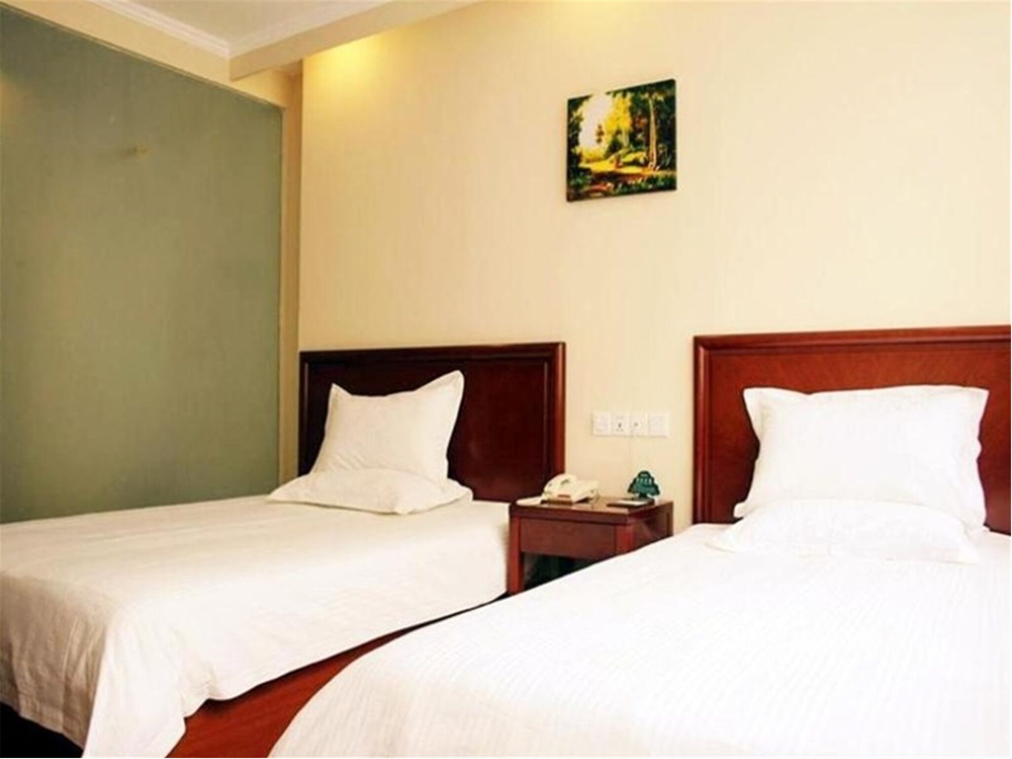 A hotel room with two single beds, a wooden headboard, a small bedside table with a phone and a green ornament, and a painting on the wall.