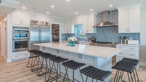 Een moderne keuken met witte kasten, een groot eiland met barstoelen, roestvrijstalen apparatuur en een spatwand met blauwe stipjes.