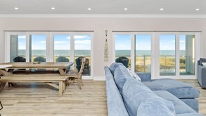 Een ruime woonkamer met een houten tafel en een blauwe bank, met grote glazen deuren die uitkijken op het strand.