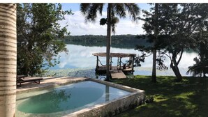 Área de piscina com deck de madeira e espreguiçadeiras, cercada por árvores e com vista para um lago tranquilo.