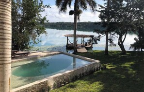 Un espace piscine doté d’une terrasse en bois et de chaises longues, entouré d’arbres et offrant une vue sur un lac paisible.
