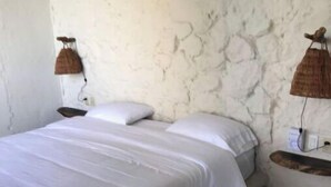 Quarto com cama, roupa de cama branca, parede de pedra e duas luminárias fixadas na parede.