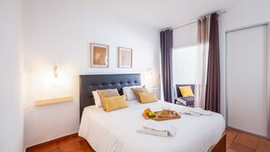 A hotel room with a bed, a tray with fruits and towels, and a chair with a cushion.