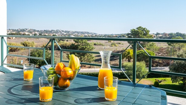 Ein Tisch mit einer Fruchtschale, zwei Orangensaftgläsern und einer Saftkanne, mit Blick auf eine Landschaft aus Häusern und Bäumen.