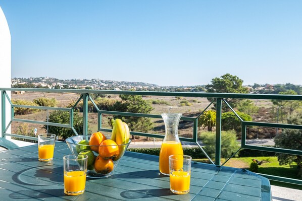 A table with a bowl of fruit and two glasses of orange juice, a pitcher of orange juice, and a view of a landscape with houses and trees.