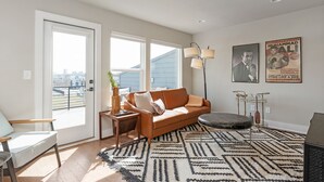 A living room with a brown sofa, a coffee table, and a patterned rug. There are two windows with blinds, a floor lamp, and a poster on the wall. - Ultimate Downtown Charlotte Oasis, 10 Guests (Charlotte)