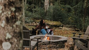 Una persona sentada junto a un hogar de piedra en un área boscosa, con sillas y una mesa de madera.