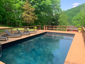 Una piscina rectangular con terraza de madera y sillones de descanso.