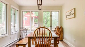 Un comedor con mesa y sillas de madera, ventanas amplias y una lámpara colgante.