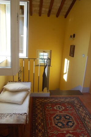 A room with yellow walls, a wooden bed with white linens, a patterned rug, and a window with a view of greenery.