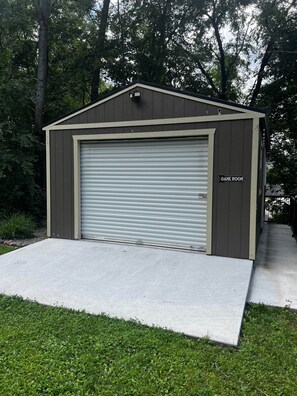 A small, single-story building with a closed garage door, labeled "GAME ROOM," surrounded by a grassy area and trees.