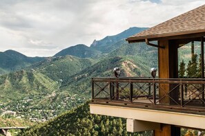 Ein hölzerner Balkon mit Metallgeländer, der auf eine gebirgige Landschaft mit einem Dorf weiter unten blickt.